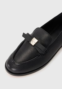 Black leather loafer with a textured finish, featuring a decorative bow and a silver accent on the upper part. Round toe and flat sole.
