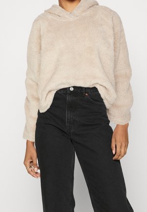 Pull à capuche beige duveteux porté avec un jean noir taille haute, recadré pour montrer le torse et les bras sans visage visible.