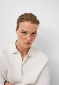 Biała koszula z kołnierzykiem z gładkiego materiału, z plisą i luźnymi rękawami. Model ma krótkie, jasnobrązowe włosy i niebieskie oczy.