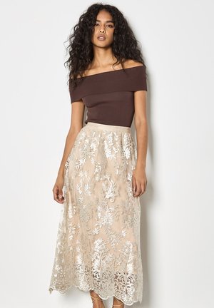 Kobieta o długich kręconych włosach, ubrana w brązową bluzkę odkrywającą ramiona i beżową koronkową spódnicę w kwiaty, stoi na tle jednolitej białej ściany.