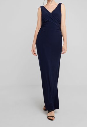 Femme portant une robe portefeuille sans manches, bleu marine, longue jusqu'au sol, avec un décolleté en V et des talons ouverts noirs, debout sur un sol gris.