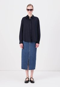 Navy button-up shirt met lange mouwen, gecombineerd met een blauwe denim maxi rok met een split aan de voorkant. Zwarte zonnebril en schoenen maken de look compleet.
