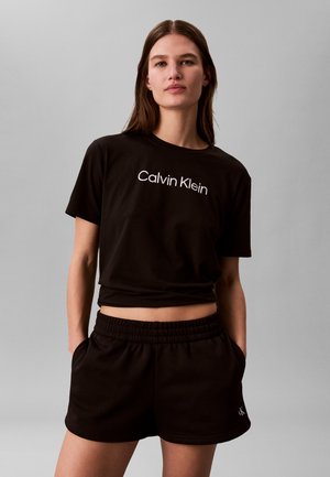 Vrouw die een zwart Calvin Klein t-shirt en bijpassende zwarte elastische taillebroek draagt, staand met handen in de zakken tegen een effen grijze achtergrond.