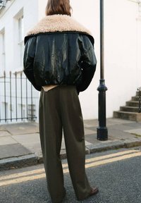Chaqueta bomber de cuero negro y brillante con un suave cuello beige, combinada con pantalones sueltos de color oliva. Los detalles visibles incluyen una tela texturizada y un corte corto.