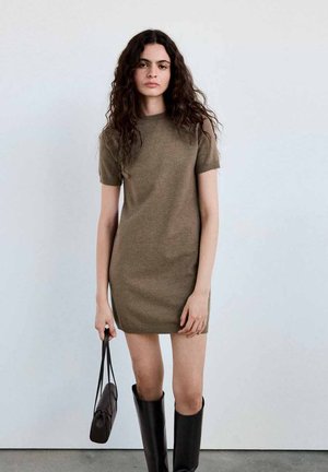 Mujer con cabello largo y rizado que lleva un vestido marrón de punto de manga corta, botas negras hasta la rodilla y sostiene un pequeño bolso negro.