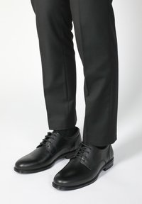 Schwarze Lederschuhe mit glatter Oberfläche, runder Spitze und Schnürdesign, kombiniert mit schwarzen Hosen und Socken.