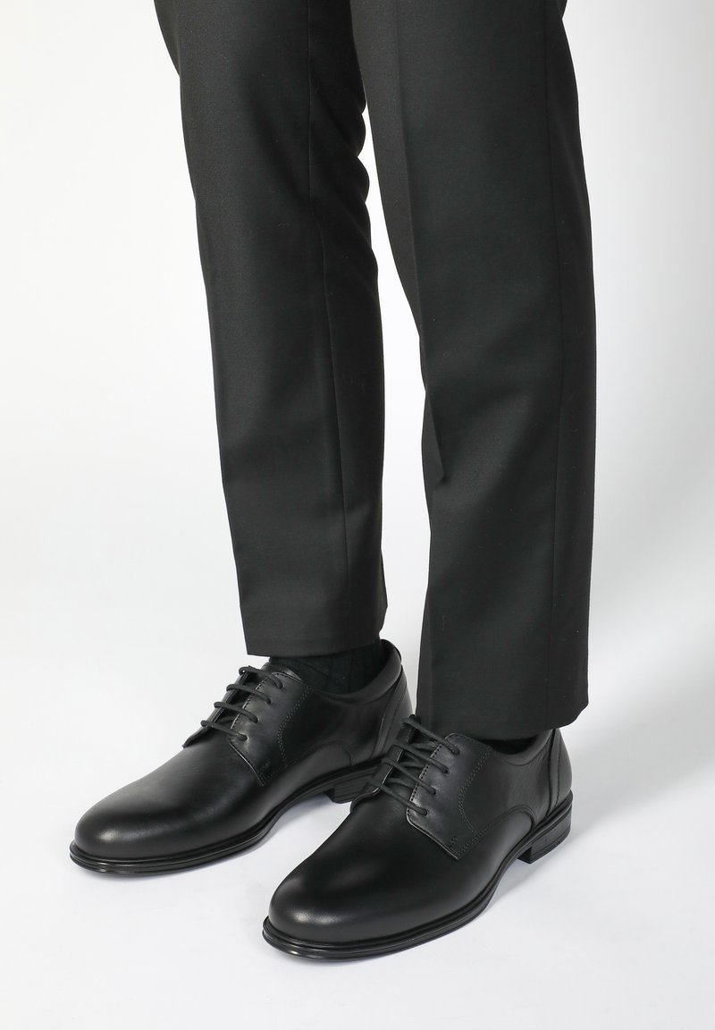 Schwarze Lederschuhe mit glatter Oberfläche, runder Spitze und Schnürdesign, kombiniert mit schwarzen Hosen und Socken.