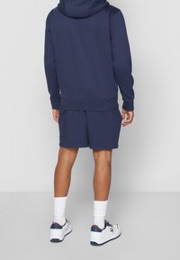 Felpa blu navy con tasca a marsupio, abbinata a pantaloni corti coordinati. Scarpe da ginnastica bianche con accenti blu navy e rossi, indossate con calzini bianchi.