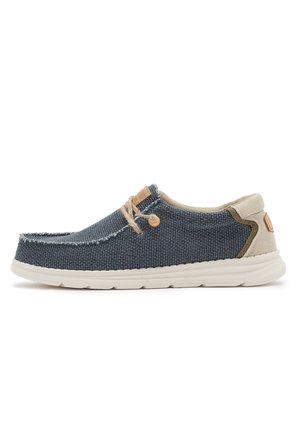 Zapatilla casual de tela azul con talón beige, suela blanca y cordones marrón claro, mostrada de lado sobre un fondo blanco.