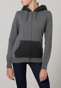 Mujer vestida con una sudadera gris con cremallera, con un bolsillo frontal texturizado en negro y forro de capucha, combinada con jeans azul oscuro, de pie frente a un fondo claro.