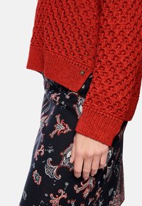 Red knit sweater with a textured pattern and ribbed cuffs, paired with a patterned skirt featuring floral designs in blue, red, and white.
