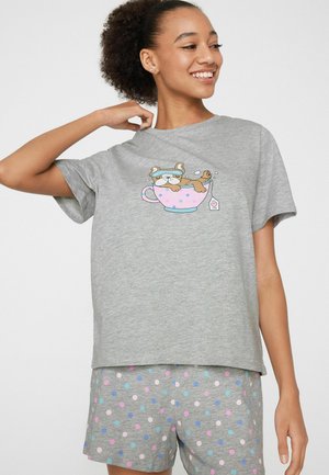 Lächelnde Frau in grauem Schlafanzug mit einem Hund, der sich auf einem Teetassen-Grafik auf dem Shirt entspannt, und bunten Punkten auf der Shorts.