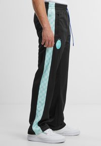 Crne sportske hlače s svijetloplavim uzorkovanim bočnim trakama. Sadrže emblema u blizini bokova i podesivi pojas s plavim vezicom.