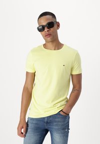 Tricou galben din bumbac, cu mâneci scurte și gât rotund, având un logo mic pe piept. Poartă jeansi albaștri, uzati.