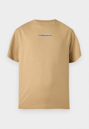 Mannelijk model staat met handen in de zakken, draagt een beige T-shirt met korte mouwen, witte shorts, witte sokken en witte sneakers tegen een effen achtergrond.