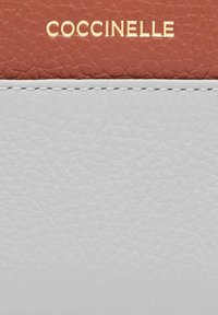 Šedá kožená peněženka s hladkým povrchem, zdobená oranžovým koženým detailem a zlatým logem "COCCINELLE" vyraženým na povrchu.