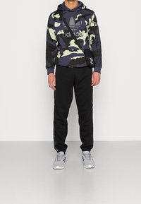 Sudadera con capucha de camuflaje en verde oscuro y azul marino con detalles grises, combinada con pantalones de chándal negros con franjas laterales y zapatillas grises.