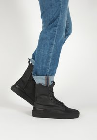 Personne portant un jean bleu retroussé et des bottines montantes noires à lacets, debout sur un fond clair.