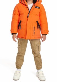 Giacca a vento arancione con dettagli neri e cerniere, abbinata a pantaloni cargo beige e sneakers bianche. Dispone di cappuccio e due tasche laterali.