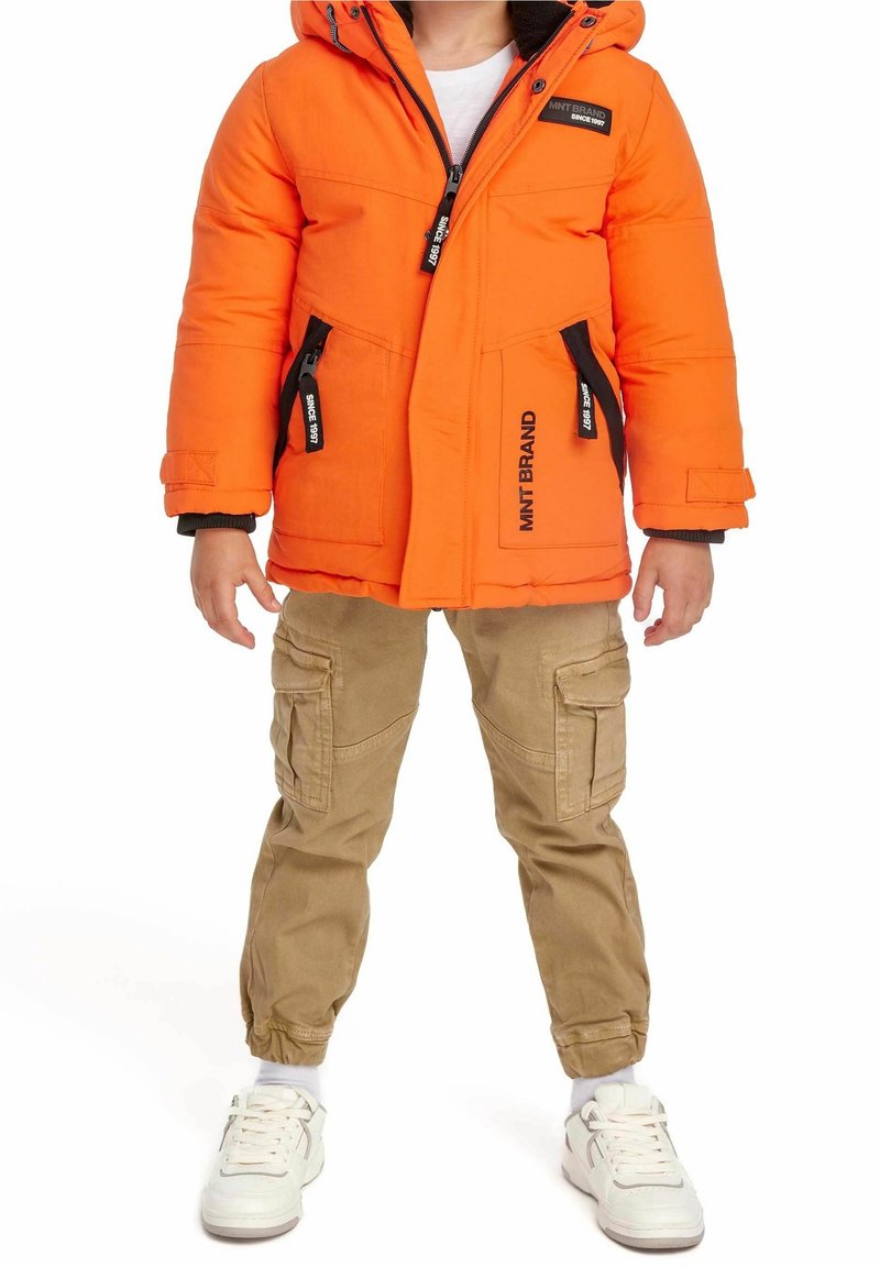Giacca a vento arancione con dettagli neri e cerniere, abbinata a pantaloni cargo beige e sneakers bianche. Dispone di cappuccio e due tasche laterali.