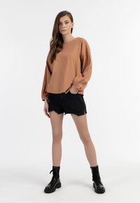 Brun langærmet top med samlede manchetter, parret med sorte slidte shorts og sorte snørelåste kampstøvler på en hvid baggrund.