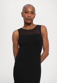 Black sleeveless dress with a sheer mesh panel at the neckline. Fitted shape, smooth texture, and subtle branding detail at the waist.