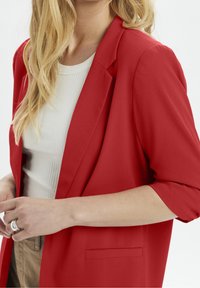 Punainen muotolöydät takki, jossa on sileä tekstuuri, lovetut läpät ja rullatut hihansuut. Sisältää yhden etutaskun ja valkoisen ribbipaidan alla.