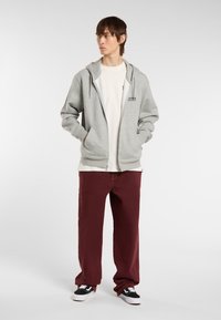 Sudadera gris con cremallera y bolsillo frontal, combinada con pantalones rectos burdeos y zapatillas negras. El modelo está de pie, con las manos en los bolsillos.