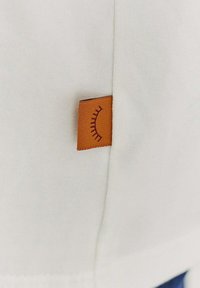 Bílá bavlněná tkanina s viditelnou oranžovou tkanou etiketou, která obsahuje minimalistický vyšívaný design. Etiketa je umístěna na postranním švu oděvu.