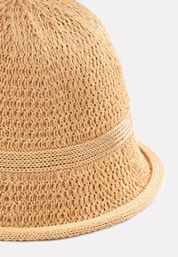 Szydełkowany kapelusz ze słomy w jasnym beżu, z teksturowanym wzorem i plecioną opaską. Posiada zaokrągloną koronę i lekko rozszerzający się brzegi.