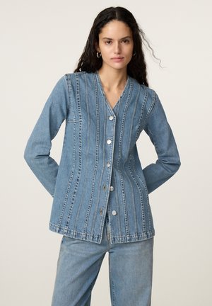 Vrouw met lang donker haar draagt een blauwe spijkerjas met knopen en verticale naden en een bijpassende spijkerbroek tegen een effen achtergrond.