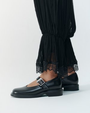Personne portant des chaussures Mary Jane en cuir noir avec boucle argentée et un pantalon noir plissé transparent avec bordure en dentelle sur fond uni.