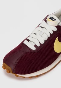 Tamsiai raudoni Nike sportbačiai su tekstūruota šenilinė viršutine dalimi, baltais raišteliais, geltonu logotipu ir rudos gumos padu su daugybe sukibimo iškyšų.