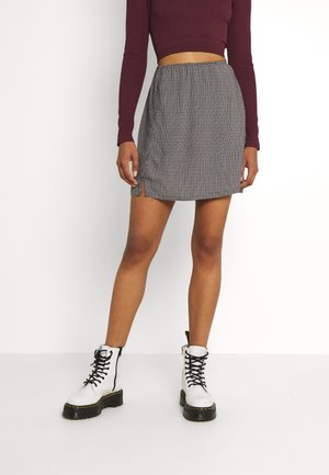 Kobieta ubrana w bordową długorękawową krótka bluzkę, czarno-białą kraciastą mini spódnicę z małymi bocznymi rozcięciami oraz białe buty wojskowe z czarnymi sznurowadłami.