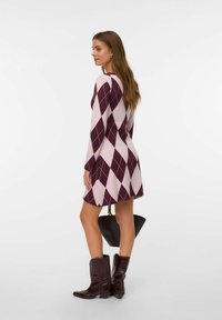 Donna che indossa un abito lungo a maniche lunghe argyle viola e rosa chiaro, stivali marroni scuri, mentre tiene una borsa nera, in piedi su uno sfondo bianco.