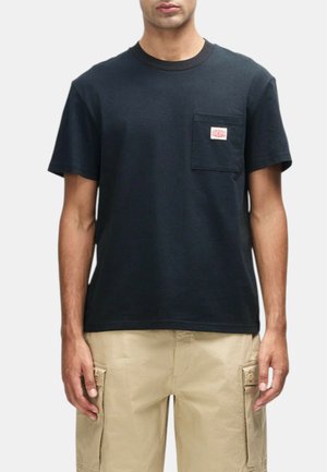 Uomo che indossa una t-shirt nera semplice a maniche corte con taschino sul petto e pantaloncini cargo beige, in piedi davanti a uno sfondo chiaro.