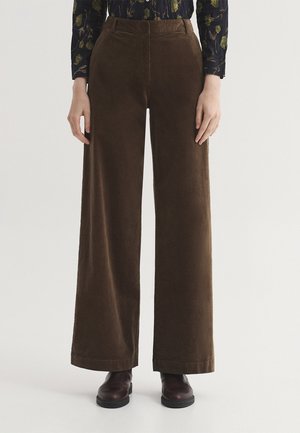 Persona con pantalones marrones de pierna ancha, zapatos marrón oscuro y una camiseta de manga larga con un patrón floral oscuro, de pie frente a un fondo blanco simple.