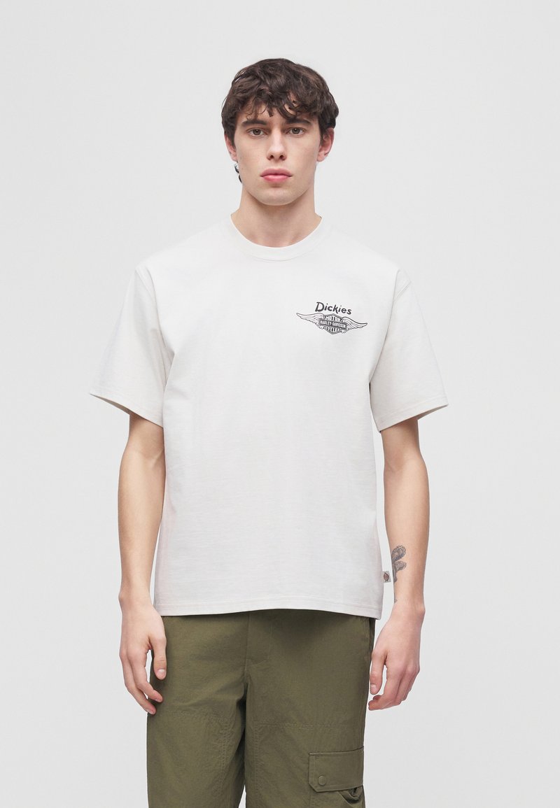 Jeune homme aux cheveux courts et foncés portant un t-shirt blanc Dickies et un pantalon cargo vert olive, debout devant un fond clair uni.