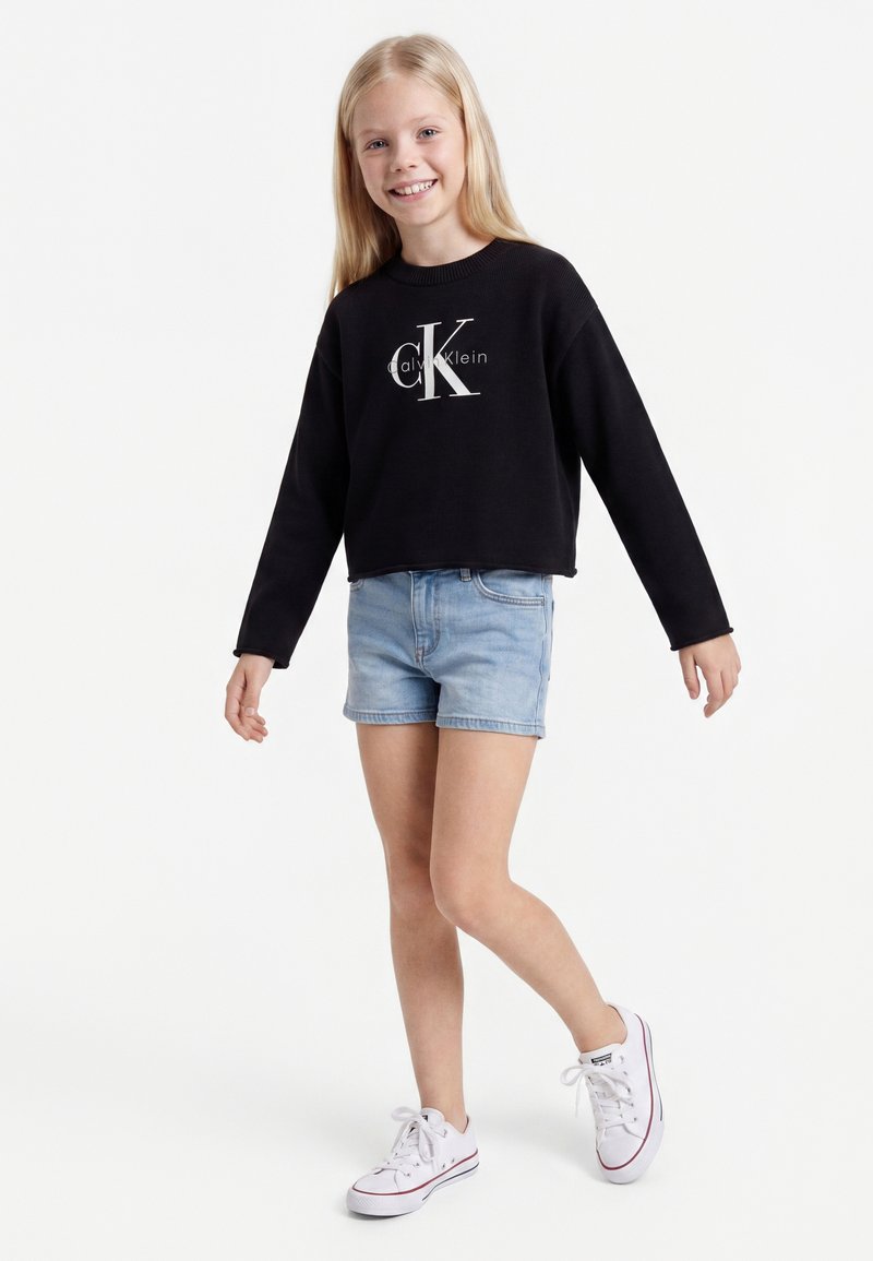 Jeune fille souriante, portant un sweat-shirt Calvin Klein noir à manches longues, un short en jean bleu clair et des baskets blanches, debout sur une jambe.