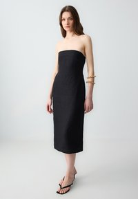 Schwarzes, trägerloses Kleid aus Leinen mit enganliegendem Oberteil und gerader Silhouette. Das Model trägt schwarze Sandalen und ein goldenes Armband am Arm.