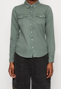 Vihreä nappikauluksellinen paita pitkillä hihoilla, jossa on kaksi rintataskua ja pyöristetty helma. Valmistettu pehmeästä kankaasta, jossa on sileä rakenne.