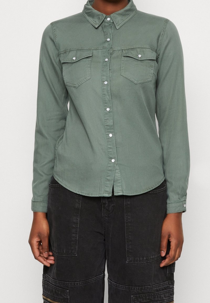 Vihreä nappikauluksellinen paita pitkillä hihoilla, jossa on kaksi rintataskua ja pyöristetty helma. Valmistettu pehmeästä kankaasta, jossa on sileä rakenne.