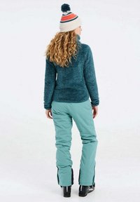 Tamsiai žalios vilnos megztinis su aukšta apykakle. Modelis vilki šviesiai žalias slidinėjimo kelnes su užtrauktukais ant kulkšnies ir juodais akcentais.