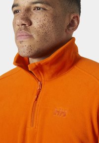 Pomarańczowy polar z pół-zamkiem, o teksturowanej powierzchni, z dyskretnym logo na klatce piersiowej i kontrastowym zamkiem.