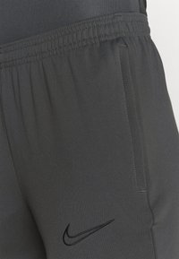 Grijze sportbroek met een gladde textuur, elastische tailleband, zijzak en een zwart Nike-logo geborduurd aan de linkerkant onderaan.
