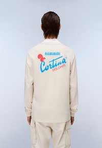 Μπεζ μπλούζα μακρύ μανίκι με μπλε και κόκκινο γραφικό σχέδιο στην πλάτη, που περιλαμβάνει το κείμενο "Cortina DOLOMITI" και λεπτομέρειες λογότυπου. Μαλακή υφή.