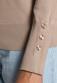 Pull beige clair avec texture côtelée et trois boutons décoratifs en or rose sur le poignet. Taille ajustée pour un look près du corps, associé à un denim.