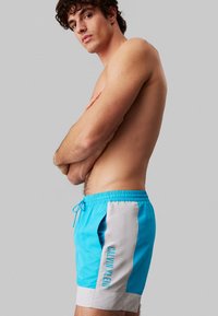 Costume da bagno da uomo in azzurro brillante con pannello laterale grigio. Caratterizzato da vita elastica, coulisse e logo stampato sul lato.