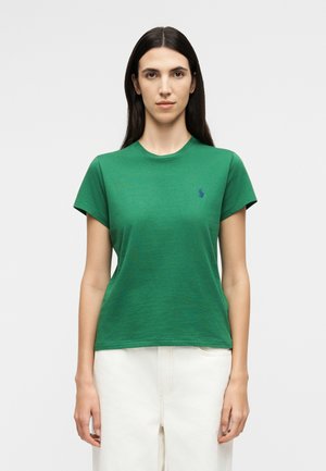 Γυναίκα με μακριά σκούρα μαλλιά φοράει απλό πράσινο μπλουζάκι με κοντά μανίκια και λευκό παντελόνι, στέκεται μπροστά σε απλό ανοιχτόφωτο φόντο.