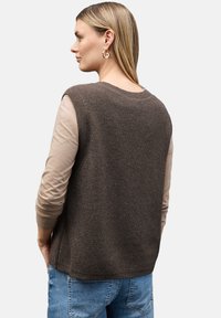 Gilet en tricot marron avec une texture côtelée, porté par-dessus une chemise à manches longues beige clair. Un jean en denim bleu visible en bas.